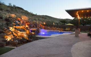 Hillside pool at dusk with a lit rock waterfall and pergola